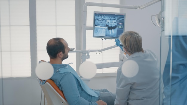 Зрелый стоматолог указывает на рентгенограмму зубов на мониторе, чтобы объяснить пациенту с зубной болью диагноз стоматитита. Стоматолог показывает результаты рентгеновского сканирования человеку с болями в полости рта.
