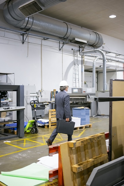 Free photo mature engineer in formal suit and hardhat walking on plant floor, carrying case with tools