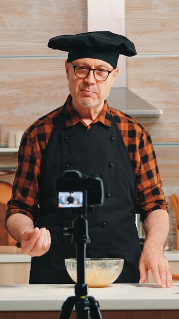 Mature experienced baker explaining recipe for audience recording tutorial for social media. Retired blogger chef influencer using internet technology communicating, shooting with digital equipment