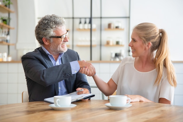 Mature expert meeting with young customer over cup of coffee at co-working, holding documents and shaking hands