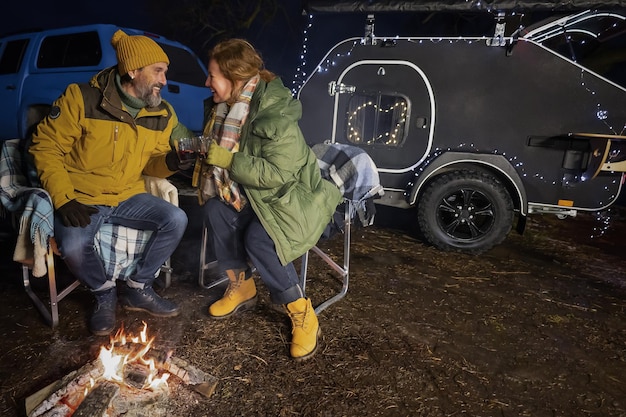 Mature family at campfire near camper away in nature Mature couple at campfire romantic dinner hot natural tea sitting on the plaid blankets in winter relaxation journey Family journey concept