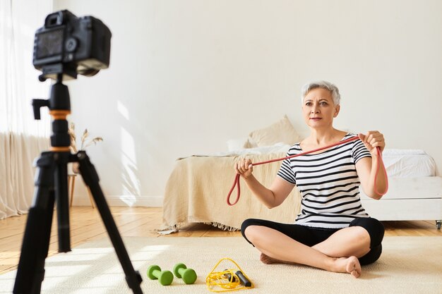 Mature female blogger sitting on floor with dumbbells and skipping rope, recording video for her blog, holding elastic band, looking  on tripod stand, explaining how to use sports equipment