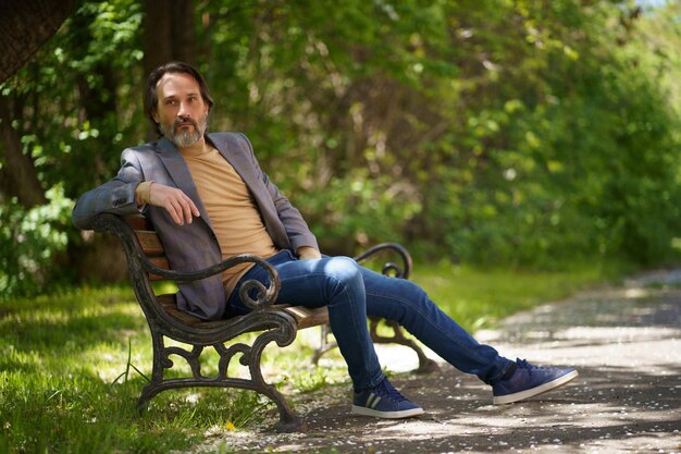 Mature freelancer man with grey beard spend time sitting on the bench at the park enjoying free time or waiting on colleagues wearing casual jacket and jeans