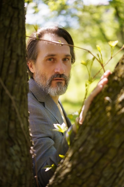 Mature grey beard businessman wearing casual grey jacket looking thru tree trunks leaning on one Life after 40 years concept problems and depression Middle age crisis