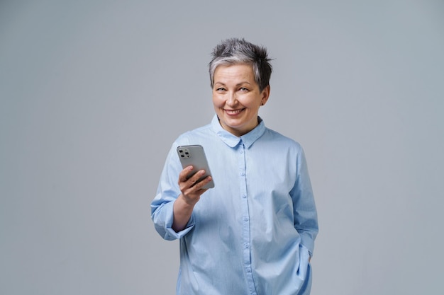 Зрелая седая деловая женщина со смартфоном в руке смотрит на камеру, делая покупки онлайн, проверяя социальные сети Красивая женщина в синей рубашке на белом фоне Скопируйте пространство