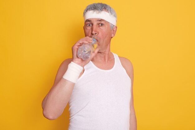 Mature grey haired male dresses white headband and sleeveless t shirt, drinking water during physical exercising, 