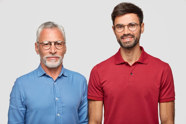 成熟した白髪の男と彼の大人の息子は白い壁に立ち、会った後の表情を喜ばせ、丸い眼鏡をかけ、1つの友好的な家族です。人と世代の概念