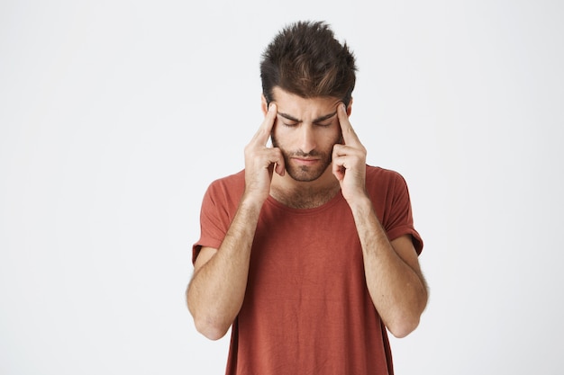 Mature italian unshaved student in red tshirt holding hands on forehead looking extremely exhausted after hard day on work. Body language.