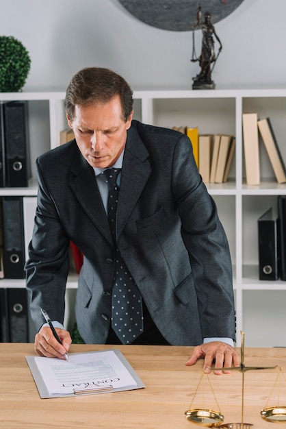 Mature male lawyer signing contract document in front of justice scale