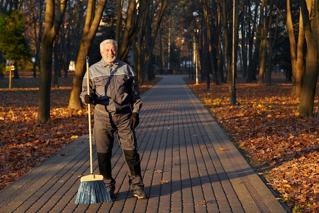 Зрелый рабочий-мужчина позирует перед камерой с метлой в парке, вид спереди на счастливого бородатого мужчину в возрасте