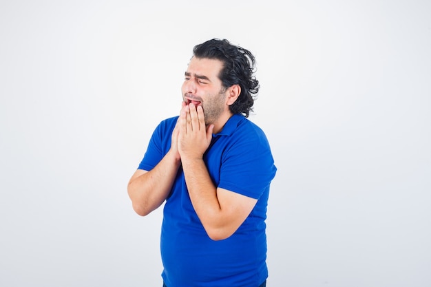 Mature man in blue t-shirt holding hands near mouth and looking mournful , front view.