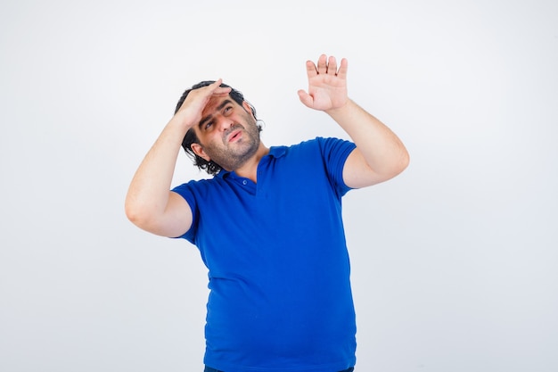 Mature man in blue t-shirt, jeans looking far away with hand over head, stretching hand and looking focused , front view.