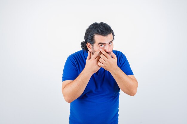 Mature man in blue t-shirt, jeans pulling down eyelid and looking serious , front view.