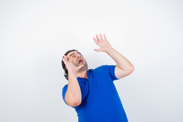 Mature man in blue t-shirt, raising hands in scared manners and looking scared, front view.