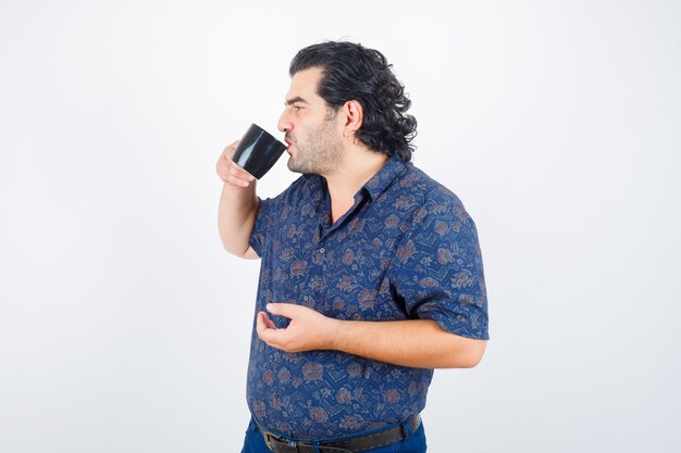 Mature man drinking while looking away in shirt and looking delighted , front view.