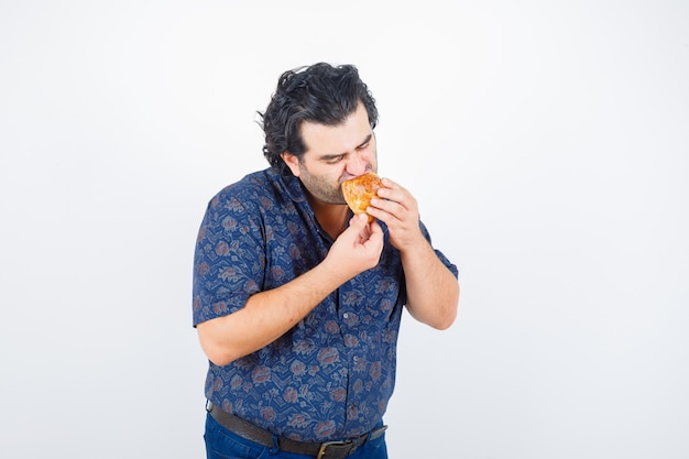 Mature man eating pastry product in shirt and looking delighted , front view.