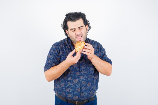 Зрелый мужчина ест кондитерские изделия в рубашке и выглядит голодным, вид спереди.