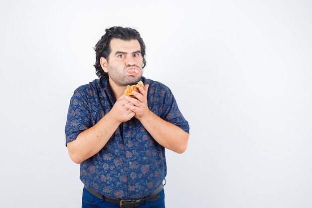 Mature man eating pastry product while looking at camera in shirt and looking serious , front view.