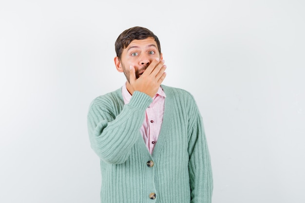 Mature man in green cardigan covering mouth with hand and looking surprised , front view.