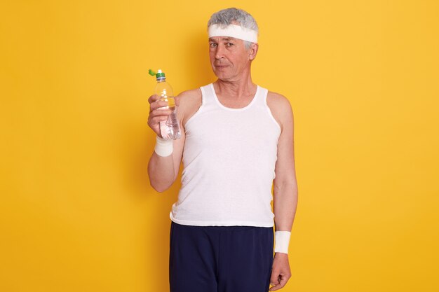 Mature man in headband and holding bottle of water, resting between sports sets, wearing t shirt and trousers