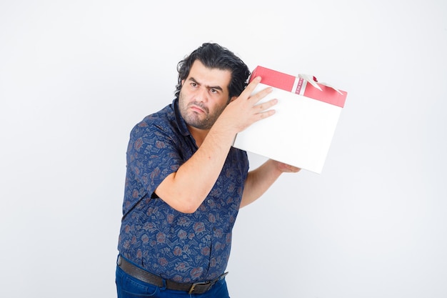 Mature man holding gift box near ear in shirt and looking curious , front view.