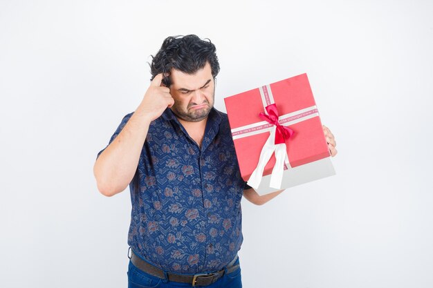 Mature man holding gift box while scratching head in shirt and looking thoughtful. front view.