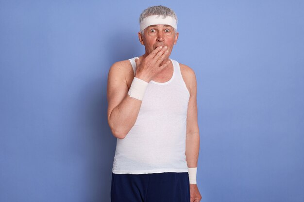 Mature man holding hands on his mouth, keeps mouth wide opened, male rejoice at unexpected news, wearing white t-shirt and hairband.