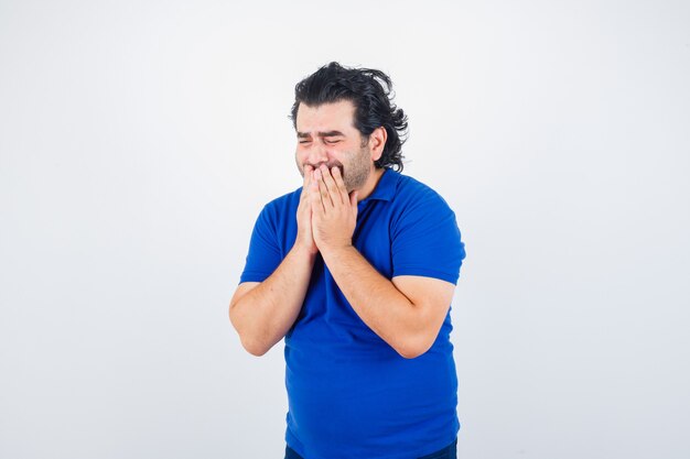 Mature man holding hands on mouth in blue t-shirt and looking mournful. front view.