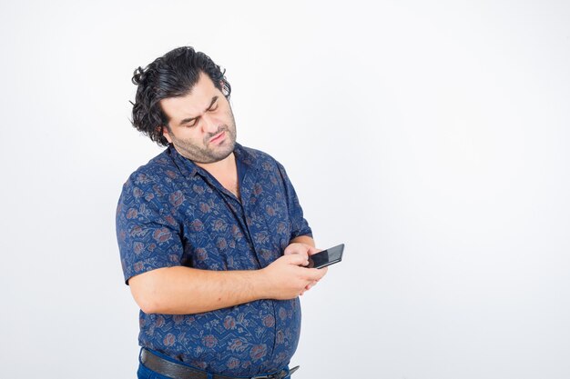 Зрелый мужчина смотрит на мобильный телефон в рубашке и смотрит задумчивый, вид спереди.