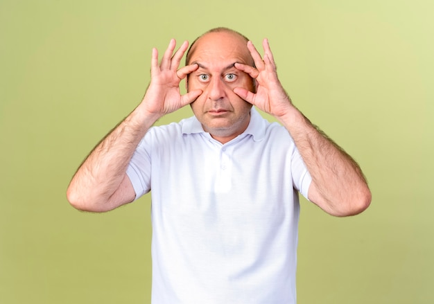 зрелый мужчина открывает глаза пальцами, изолированными на оливково-зеленой стене