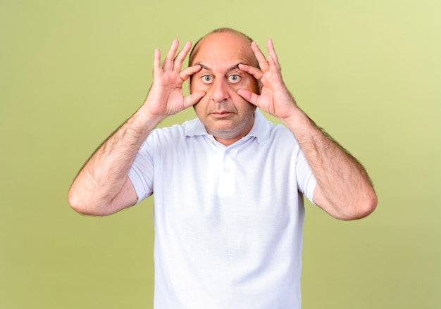  mature man opening eyes with  fingers isolated on olive green wall