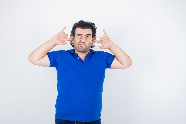 Mature man plugging ears with fingers, curving lips in blue t-shirt and looking pensive . front view.