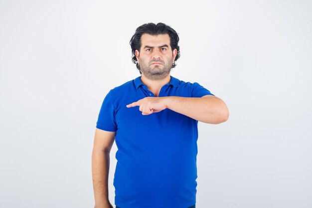 Mature man pointing left with index finger in blue t-shirt, looking angry. front view.