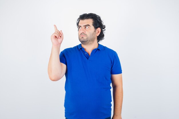 Зрелый мужчина указывает влево указательным пальцем в синей футболке, джинсах и выглядит сосредоточенным. передний план.