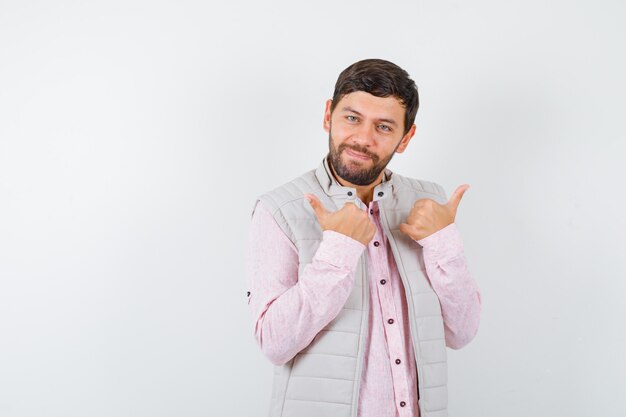 Mature man pointing to opposite directions with thumbs in shirt, sleeveless jacket and looking cheerful. front view.