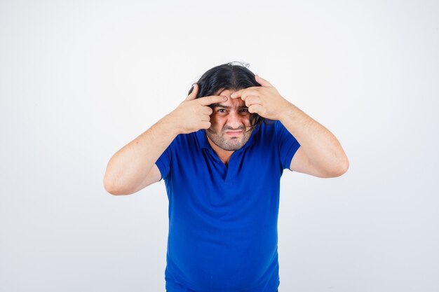 Mature man popping pimple in blue t-shirt, jeans and looking focused. front view.