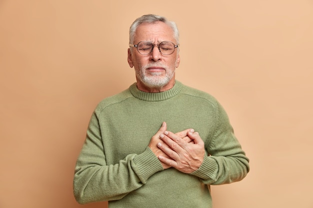 Mature man presses hands to heart has cardio problems suffers heart attack and chest pain stands with closed eyes needs doctors help wears casual jumper isolated over brown wall