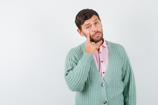 Mature man pulling eyelid down in shirt, cardigan and looking upset. front view.