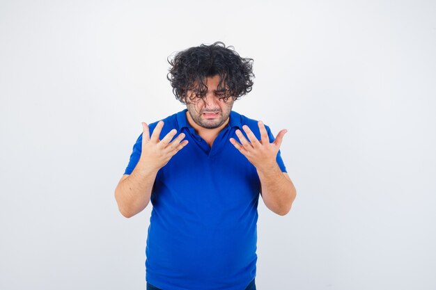 Mature man raising hands in puzzled gesture in blue t-shirt and looking wistful , front view.