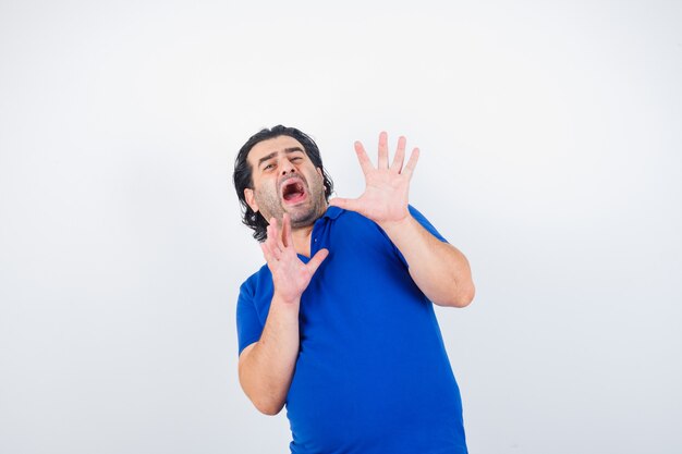 Mature man raising hands to stop in blue t-shirt and looking scared, front view.