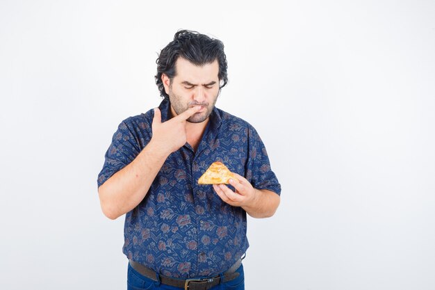 Mature man in shirt looking at pastry product while holding finger on mouth and looking thoughtful , front view.