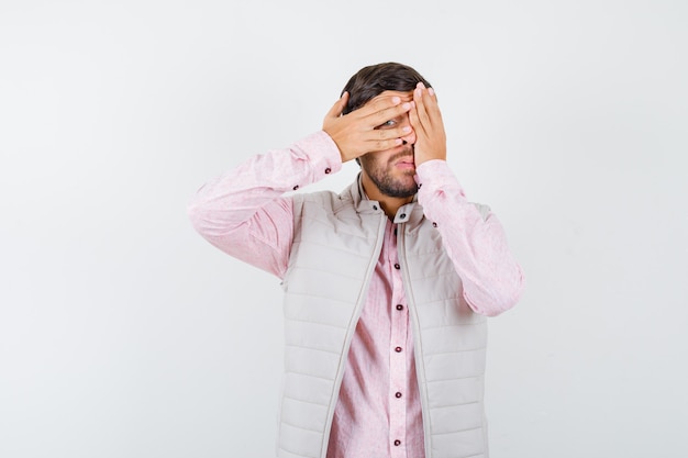 Uomo maturo in camicia, giacca senza maniche guardando attraverso le dita e guardando curioso, vista frontale.