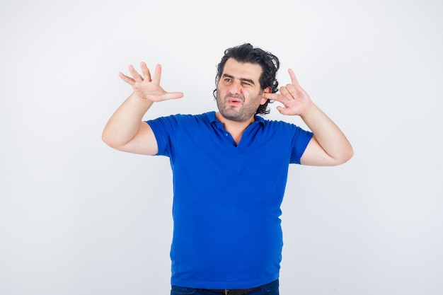 Mature man showing stop gesture, plugging ear with finger in blue t-shirt and looking bored. front view.