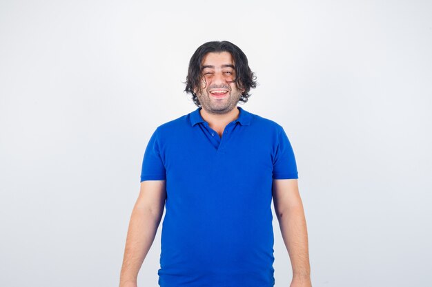 まっすぐ立っている成熟した男、青いTシャツ、ジーンズに顔をゆがめ、陽気に見える、正面図。