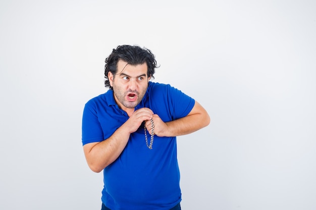 Mature man in t-shirt looking away while holding hands near chest and looking scared , front view.