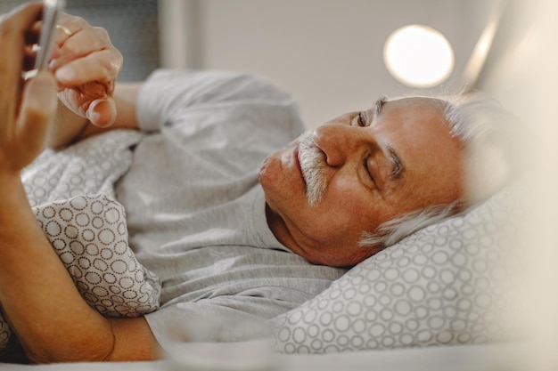 Зрелый мужчина обменивается текстовыми сообщениями на мобильном телефоне, лежа в постели