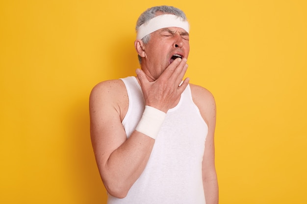 Mature man wearing white clothing covering mouth with his palm and yawning, standing against yellow wall, looks sleepy and tired