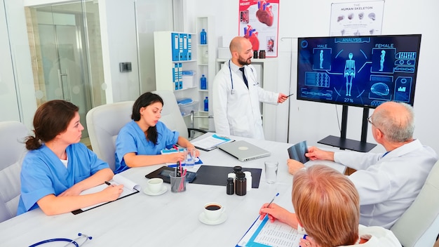 Mature medical physician explaining treatment to nurses during healthcare seminar pointing at digital monitor. Clinic herapist discussing with colleagues about disease, medicine professional.
