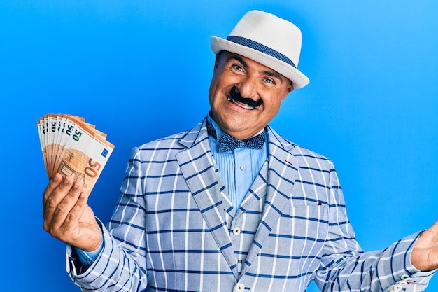 Mature middle east man with mustache wearing elegant vintage style holding euros banknotes celebrating achievement with happy smile and winner expression with raised hand