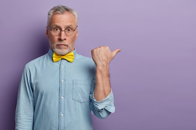 Free photo mature old man with grey hair and beard points thumb aside, has surprised face expression, wears big round glasses